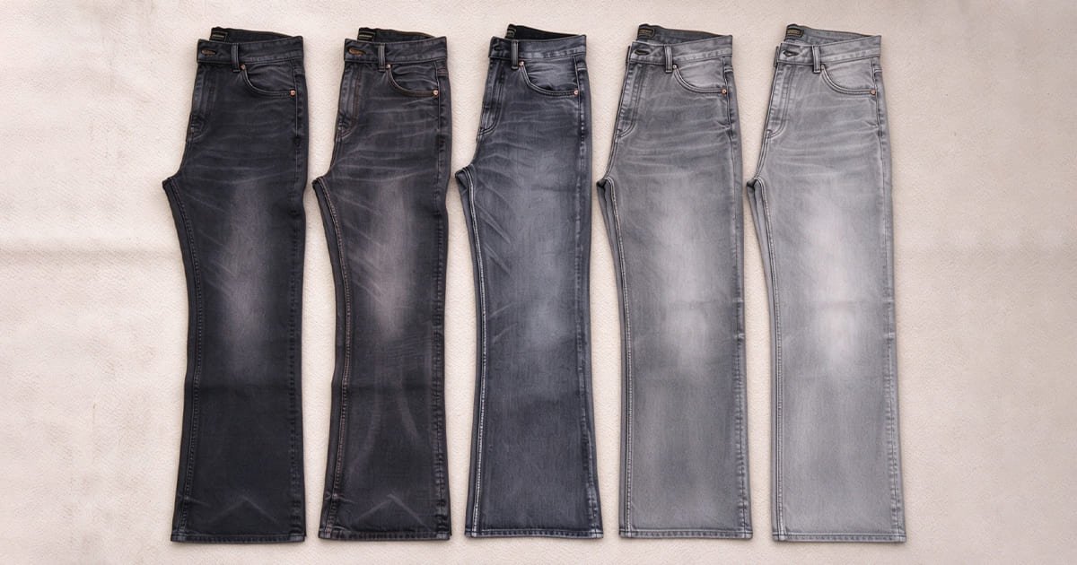 Top-down flat lay shot of five pairs of jeans arranged left to right from deep black to pale grey, showcasing a spectrum of fading wash results for black denim